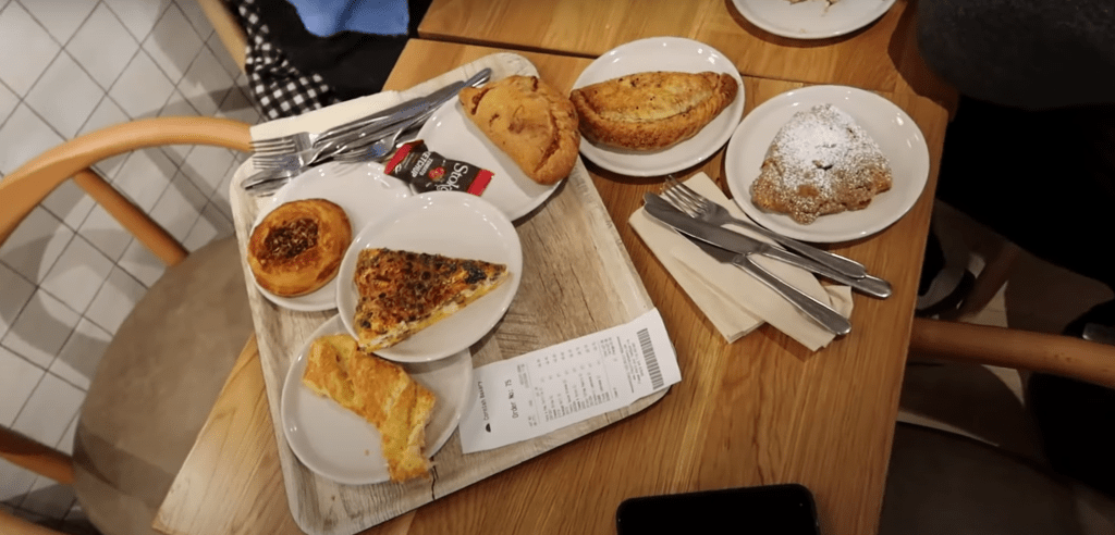 A wooden tray filled with a selection of baked goods, including quiche, cheese straws, danishes, and pasties, shared by Alfie, Zoe, Poppy, and Sean.