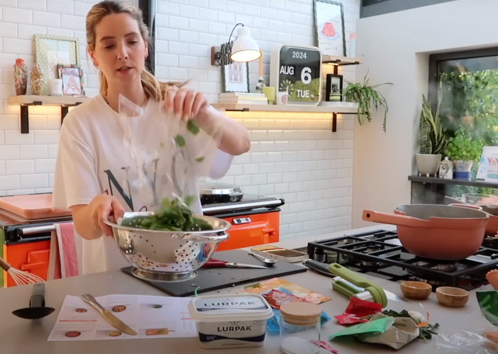 Zoe Sugg preparing HelloFresh ingredients in her bright kitchen, pouring fresh spinach into a colander while making Cajun-Spiced Rigatoni