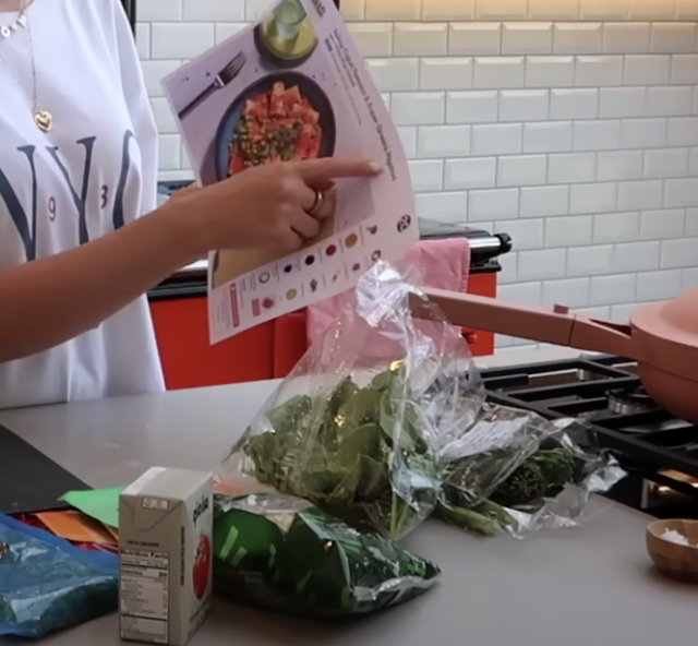 Zoe Sugg pointing at a HelloFresh recipe card for Cajun-Spiced Tomato & Super Greens Rigatoni while cooking in her modern home kitchen.